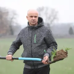 Mężczyzna w szarej bluzie z kapturem trzyma łopatę z bryłą ziemi i młodymi roślinami. Może to być sadzonka jęczmienia.