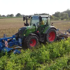Zielony traktor z maszynami rolniczymi pracuje na polu, za nim widać pole i zieleń.