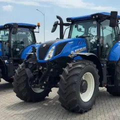Dwa niebieskie ciągniki New Holland stoją obok siebie na placu. Jeden z nich jest na pierwszym planie, z widocznymi dużymi oponami i kabiną.