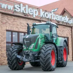 Potężny zielony traktor Fendt 936 z czerwonymi felgami stoi przed budynkiem sklepu.