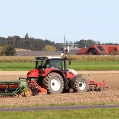 Czerwony traktor z siewnikiem na polu, w tle domy i las.