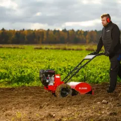 Mężczyzna w słuchawkach ochronnych pracuje glebogryzarką, spulchniając ziemię na polu. Glebogryzarka służy do przygotowania gleby pod uprawę.