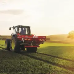 Traktor z rozsiewaczem na polu, przygotowuje glebę pod zasiewy. Słońce zachodzi, rzucając długie cienie. To idealny czas na nawożenie, by zapewnić zdrowy wzrost roślin i obfite plony, w tym kukurydzy i innych zbóż.