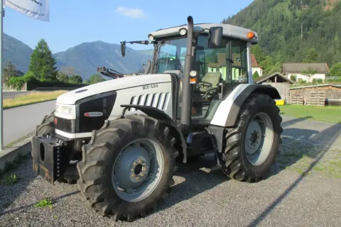 Biały traktor Hurlimann XT-130 stoi na żwirowej drodze, w tle góry i domy.
