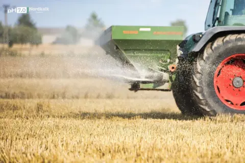 Traktor rozsiewa wapno magnezowe na polu. Drobinki nawozu unoszą się w powietrzu, poprawiając jakość gleby.