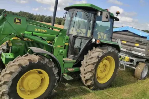 Zielony traktor John Deere 2140 z ładowaczem czołowym i przyczepą na tle łąki i lasu.