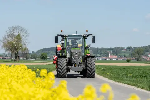 Zielony traktor Fendt 516 jedzie drogą przez kwitnące pola rzepaku, w tle widać wieś.