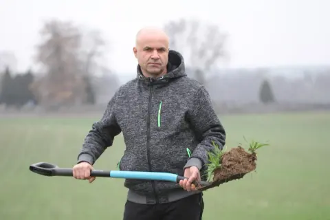 Mężczyzna w szarej bluzie z kapturem trzyma łopatę z bryłą ziemi i młodymi roślinami. Może to być sadzonka jęczmienia.