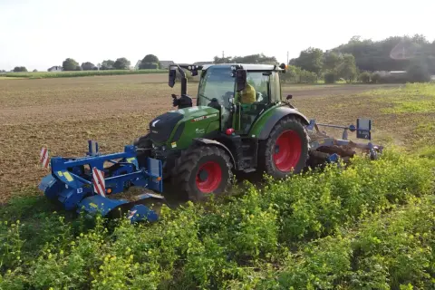 Zielony traktor z maszynami rolniczymi pracuje na polu, za nim widać pole i zieleń.
