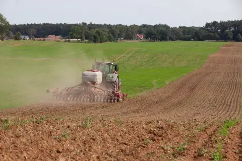 Traktor z siewnikiem HORSCH rozsiewa nawóz, prawdopodobnie fosforan, na polu. W tle las i domy.
