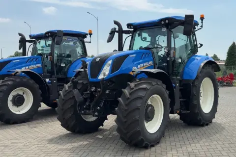 Dwa niebieskie ciągniki New Holland stoją obok siebie na placu. Jeden z nich jest na pierwszym planie, z widocznymi dużymi oponami i kabiną.