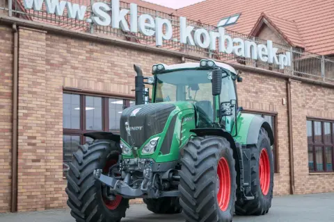 Potężny zielony traktor Fendt 936 z czerwonymi felgami stoi przed budynkiem sklepu.
