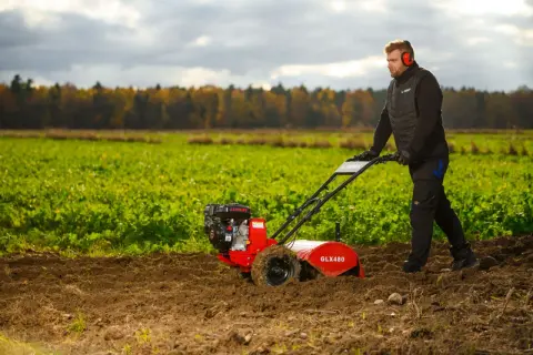 Mężczyzna w słuchawkach ochronnych pracuje glebogryzarką, spulchniając ziemię na polu. Glebogryzarka służy do przygotowania gleby pod uprawę.