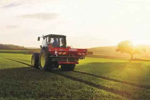 Traktor z rozsiewaczem na polu, przygotowuje glebę pod zasiewy. Słońce zachodzi, rzucając długie cienie. To idealny czas na nawożenie, by zapewnić zdrowy wzrost roślin i obfite plony, w tym kukurydzy i innych zbóż.