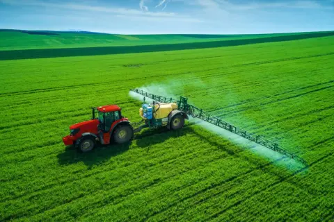 Czerwony traktor z opryskiwaczem na zielonym polu, rozpyla nawóz, by zapewnić roślinom saletrę.