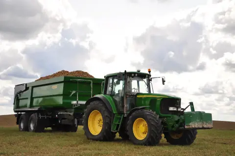 Zielony traktor John Deere z przyczepą pełną ziemniaków, wyposażoną w hamulec najazdowy, gotowy do pracy na polu.