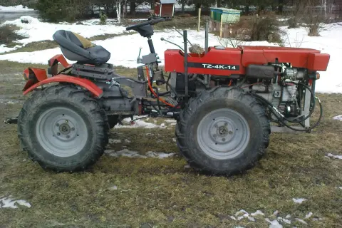 Czerwony traktor TZ-4K-14 z dużymi kołami stoi na trawiastym podwórku, częściowo pokrytym śniegiem.