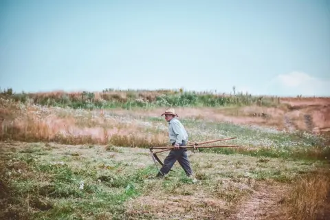 Mężczyzna w kapeluszu niesie kosę przez łąkę. Delikatna mżawka otula krajobraz, tworząc mglistą atmosferę.