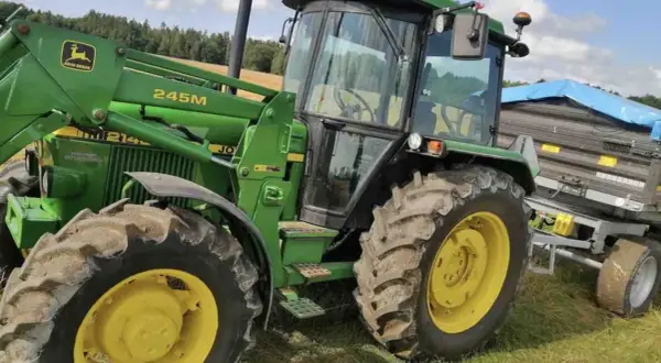 Zielony traktor John Deere 2140 z ładowaczem czołowym i przyczepą na tle łąki i lasu.
