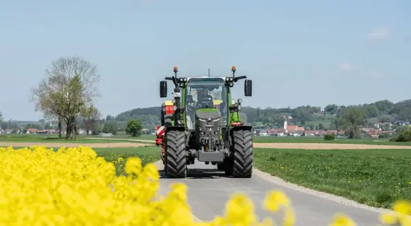 Zielony traktor Fendt 516 jedzie drogą przez kwitnące pola rzepaku, w tle widać wieś.