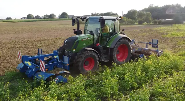 Zielony traktor z maszynami rolniczymi pracuje na polu, za nim widać pole i zieleń.