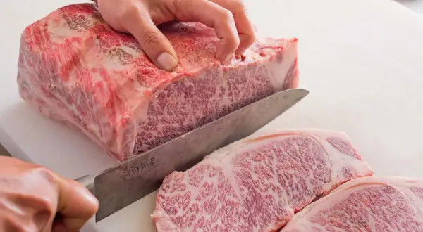 Ręka kroi luksusowe mięso wagyu na desce. Widoczne marmurkowe przerosty tłuszczu świadczą o jego wyjątkowej jakości.