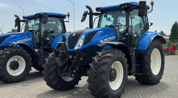 Dwa niebieskie ciągniki New Holland stoją obok siebie na placu. Jeden z nich jest na pierwszym planie, z widocznymi dużymi oponami i kabiną.