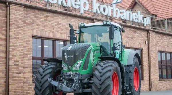 Potężny zielony traktor Fendt 936 z czerwonymi felgami stoi przed budynkiem sklepu.