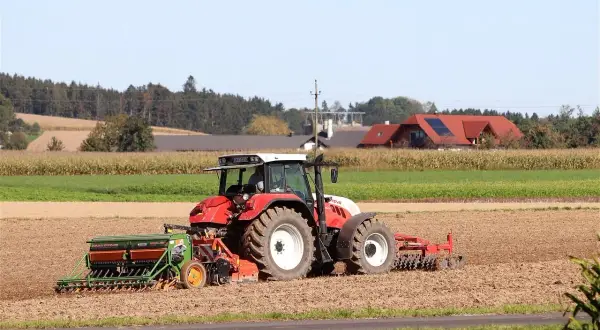 Czerwony traktor z siewnikiem na polu, w tle domy i las.