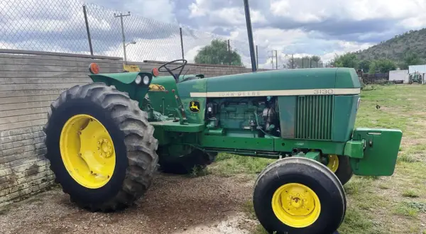 Zielony traktor John Deere 3130 z żółtymi felgami stoi na żwirowym podwórzu, gotowy do pracy na polu.