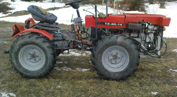 Czerwony traktor TZ-4K-14 z dużymi kołami stoi na trawiastym podwórku, częściowo pokrytym śniegiem.