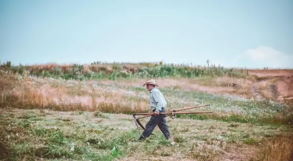 Mężczyzna w kapeluszu niesie kosę przez łąkę. Delikatna mżawka otula krajobraz, tworząc mglistą atmosferę.