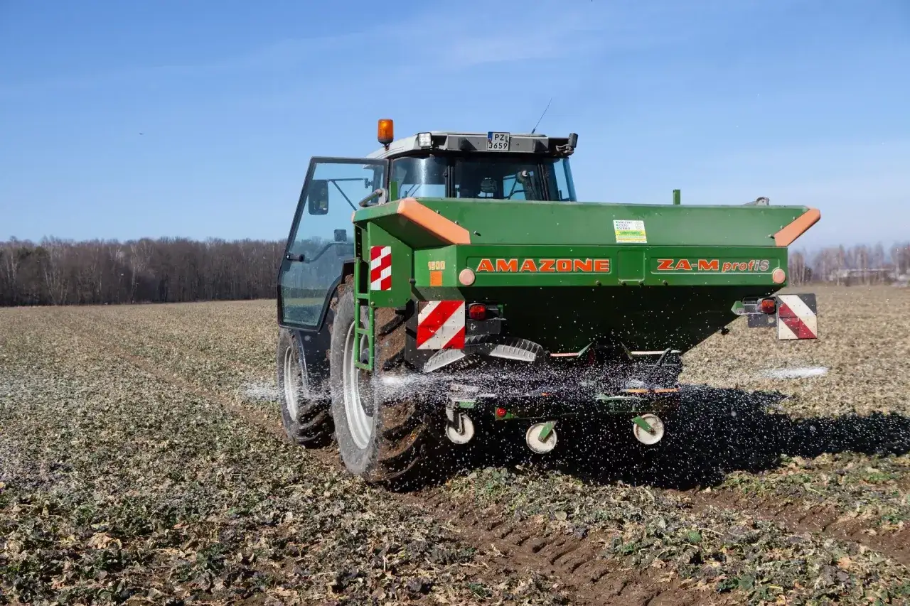 Traktor z rozsiewaczem Amazone ZA-M profiS na polu, rozprowadzający nawóz, być może nitromag, dla lepszego wzrostu roślin.
