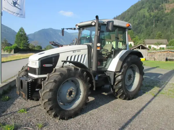 Biały traktor Hurlimann XT-130 stoi na żwirowej drodze, w tle góry i domy.