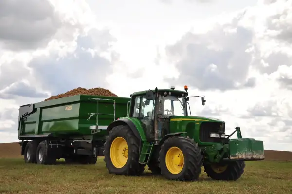 Zielony traktor John Deere z przyczepą pełną ziemniaków, wyposażoną w hamulec najazdowy, gotowy do pracy na polu.