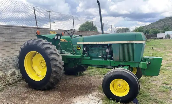 Zielony traktor John Deere 3130 z żółtymi felgami stoi na żwirowym podwórzu, gotowy do pracy na polu.