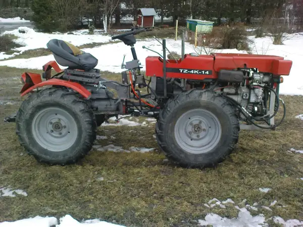 Czerwony traktor TZ-4K-14 z dużymi kołami stoi na trawiastym podwórku, częściowo pokrytym śniegiem.