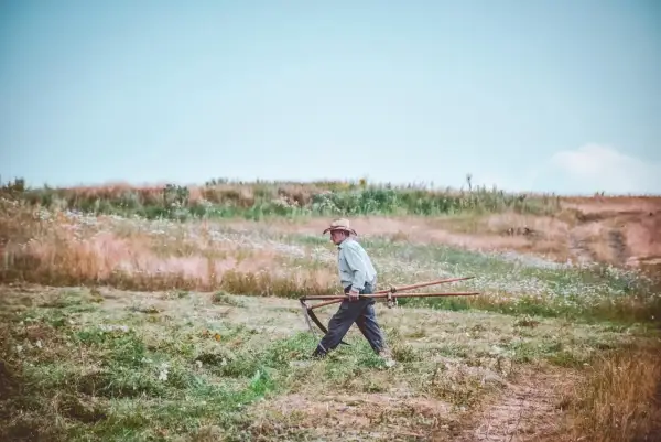 Mężczyzna w kapeluszu niesie kosę przez łąkę. Delikatna mżawka otula krajobraz, tworząc mglistą atmosferę.
